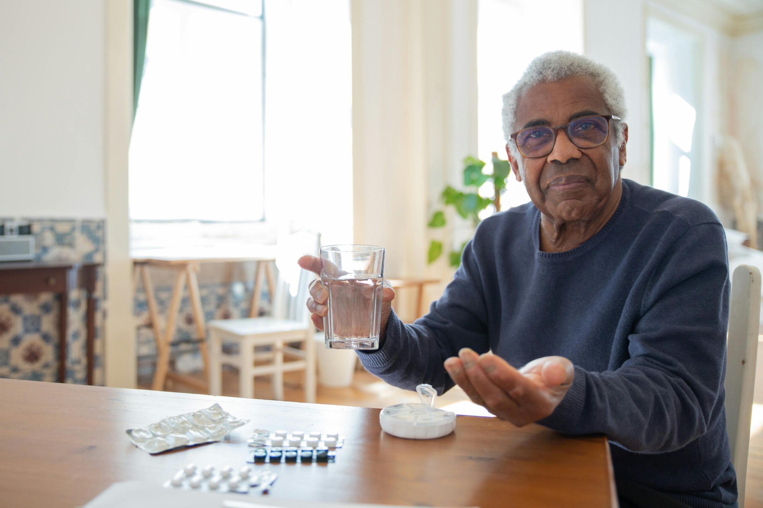 Elderly man sitting at a table at home, holding a glass of water and medication, emphasizing healthcare routine.